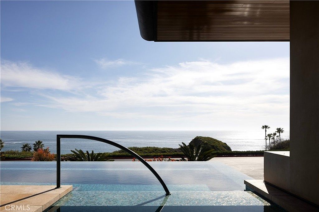 This image showcases a luxurious infinity pool overlooking the ocean. The pool features a modern design with tiled steps and a sleek black handrail. The view extends to the horizon, with glimpses of lush greenery and palm trees along the coastline, creating a serene and upscale ambiance.