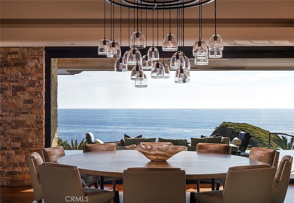 This is an interior shot of a dining room with a large round table and upholstered chairs. A modern chandelier hangs above the table, and a large window offers a stunning ocean view. The room is well-lit and appears luxurious, creating an inviting atmosphere for dining and entertaining.