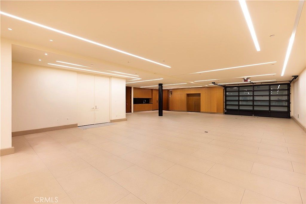 This is a spacious, modern living room with a minimalist design. The room features light-colored tile flooring, recessed lighting, and a large garage door-style window. There are built-in cabinets and a dark support column, creating a clean and open atmosphere.