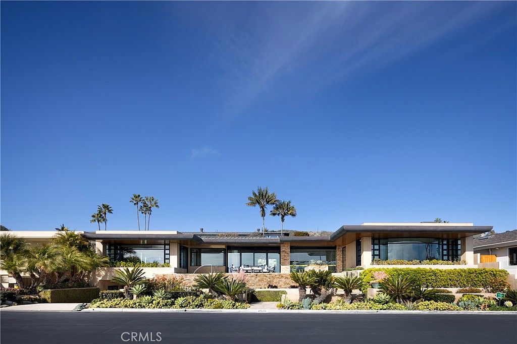 This is a front view of a modern, single-story home with a flat roof and large windows. The house is surrounded by lush landscaping, including palm trees and manicured shrubs, creating a sense of privacy and curb appeal. The clear blue sky enhances the overall impression of a well-maintained and desirable property.