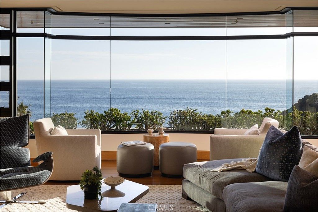 This is an interior shot of a luxurious living room with a stunning ocean view through expansive windows. The room features comfortable seating arrangements, including armchairs and a sectional sofa, complemented by stylish ottomans and a modern coffee table. The overall aesthetic is elegant and serene, emphasizing the connection to the natural surroundings.