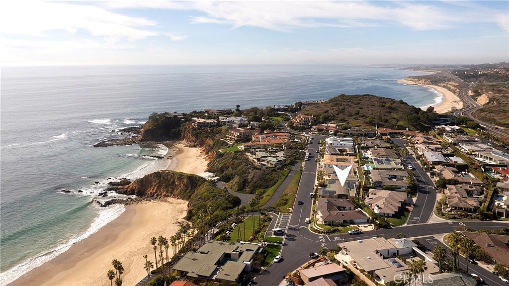 This aerial view showcases a coastal neighborhood with luxury homes, a sandy beach, and the Pacific Ocean. The homes feature well-maintained landscaping and a mix of architectural styles, creating an upscale and desirable living environment. The image highlights the proximity to the beach and the overall scenic beauty of the location.