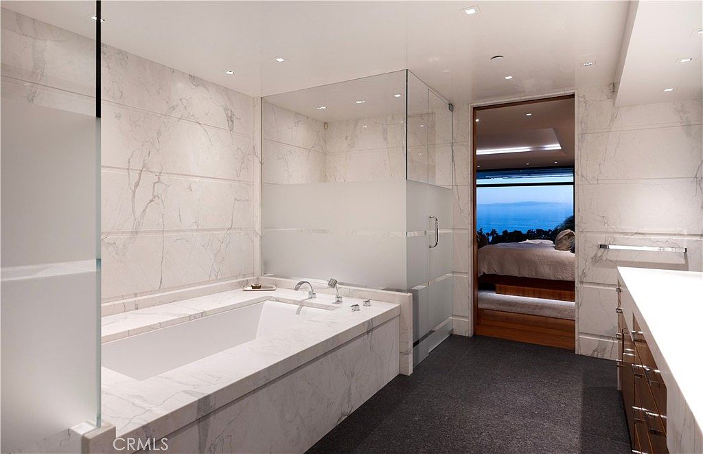 This is a luxurious primary bathroom featuring marble walls and flooring, a large soaking tub with marble surround, and a glass-enclosed shower. The bathroom opens into a bedroom with a stunning ocean view, creating a spa-like atmosphere. The design is modern and elegant, emphasizing clean lines and high-end finishes.