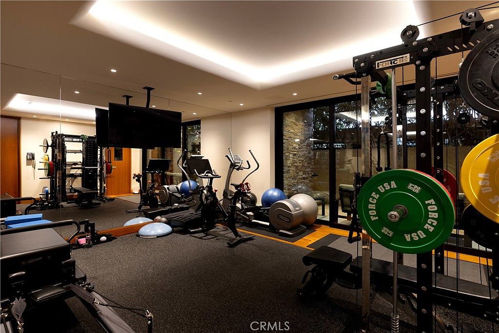 This is a well-equipped home gym featuring a variety of exercise machines, including stationary bikes, a weight rack with plates, and a rowing machine. The room has a dark rubber floor and mirrored walls, enhancing the sense of space. A large television is mounted on the wall, and natural light enters through a sliding glass door.
