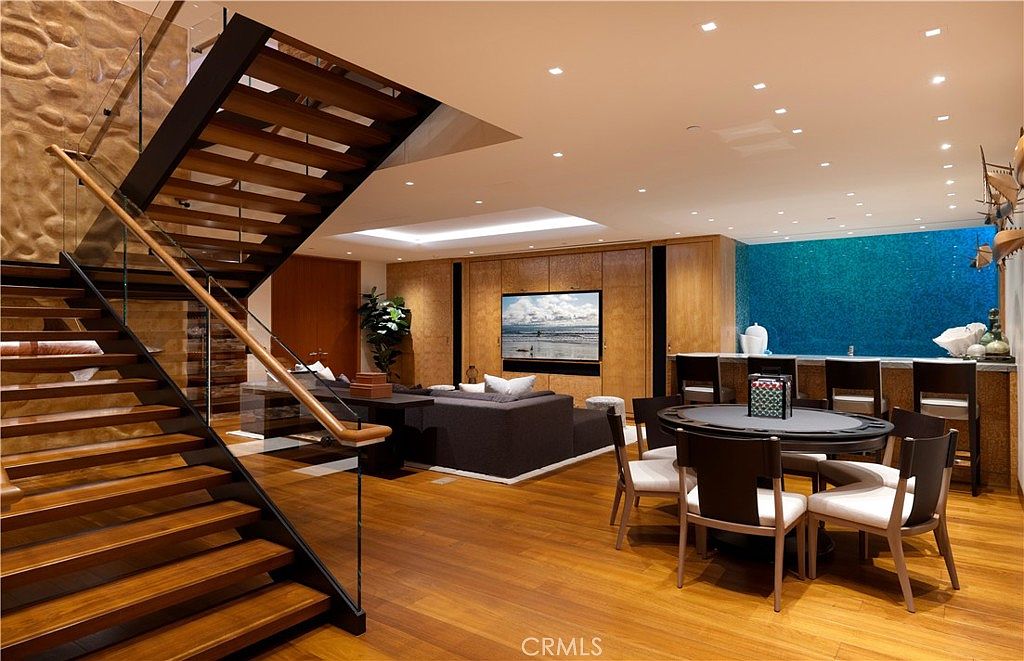 This interior shot showcases a modern living room with hardwood floors, a staircase, and a media center. The room features a dark gray sectional sofa, a poker table with chairs, and a bar area with a blue tiled wall. The overall impression is luxurious and well-designed, perfect for entertaining.