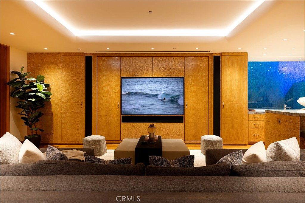 This is an interior shot of a living room featuring a large built-in entertainment center with wooden cabinetry and a flat-screen TV displaying an ocean scene. A comfortable sofa with decorative pillows sits in the foreground, facing a central ottoman and two small stools. The room is well-lit with recessed lighting, creating a warm and inviting atmosphere.