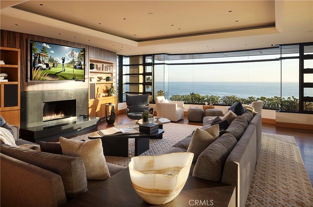 This is a luxurious living room featuring a large sectional sofa, a fireplace with a TV above it, and floor-to-ceiling windows offering stunning ocean views. The room is decorated in neutral tones with wood accents, creating a warm and inviting atmosphere. The space is well-lit and appears spacious, perfect for relaxation and entertaining.