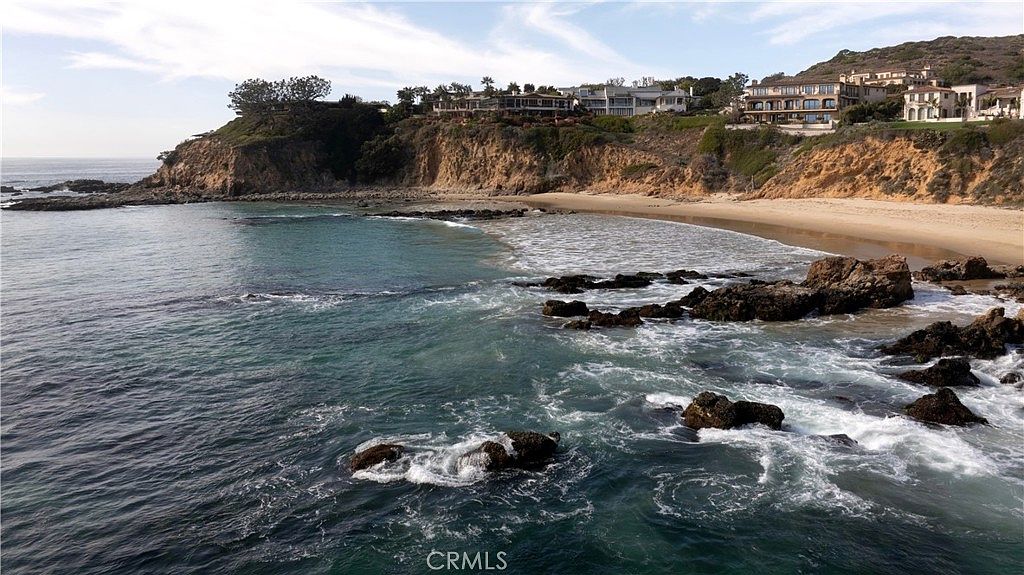 This aerial view showcases a stunning coastal property with multiple houses perched atop a cliff overlooking a picturesque beach. The turquoise water contrasts beautifully with the sandy shore and rocky outcrops, creating a serene and luxurious atmosphere. The scene emphasizes the property's prime location and breathtaking ocean views, highlighting its exclusivity and appeal.