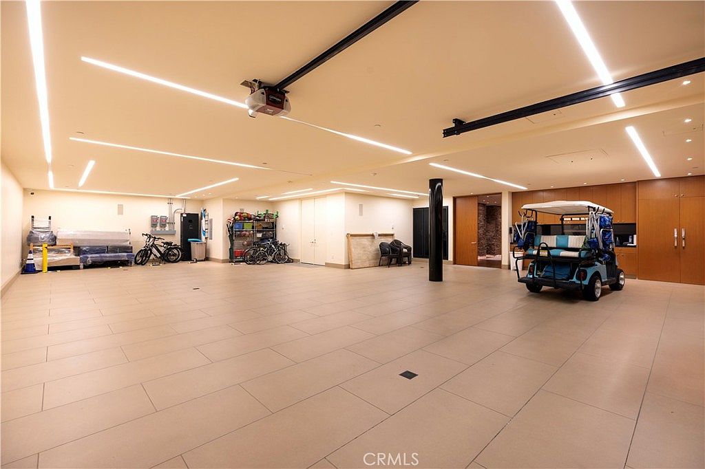 This is a spacious and well-lit garage featuring a light tile floor and recessed lighting. The garage includes storage shelves, bicycles, a golf cart, and a sofa. The walls are a combination of painted surfaces and wood paneling, creating a clean and organized space.