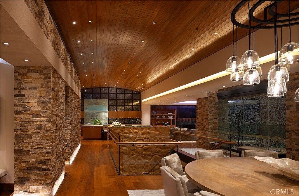 This is an interior shot of a dining room featuring a large, round wooden table with modern chairs. A unique, multi-bulb pendant light fixture hangs above the table, adding a touch of elegance. The room is decorated with stone accent walls and a wooden ceiling with recessed lighting, creating a warm and inviting atmosphere.