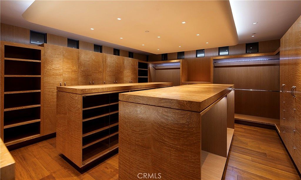 This is a luxurious walk-in closet featuring custom wood cabinetry and shelving. The closet includes two central islands with drawers and countertops, providing ample storage and display space. Recessed lighting illuminates the space, highlighting the rich wood tones and creating a warm, inviting atmosphere.