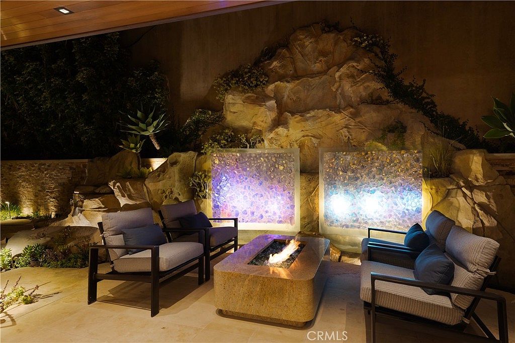 This is a beautifully designed outdoor patio area featuring comfortable seating around a modern fire pit. The backdrop includes a rock feature with integrated lighting and water elements, creating a serene and luxurious ambiance. The space is perfect for relaxation and entertaining.