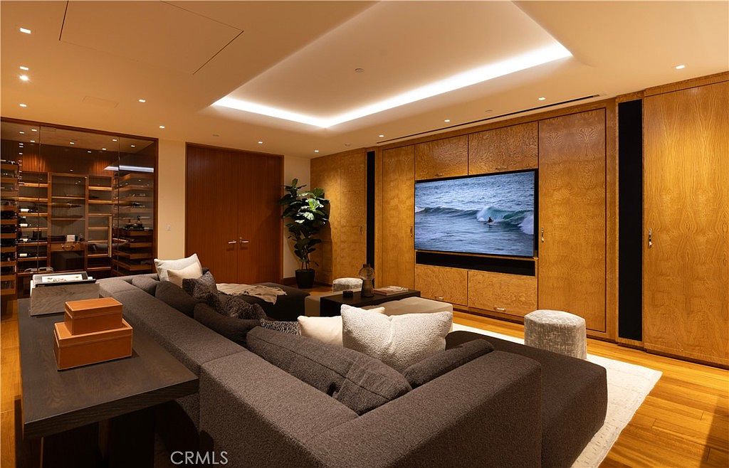 This is an interior shot of a modern living room. The room features a large sectional sofa, a wooden floor, and a built-in entertainment center with a large screen TV. The room is well-lit with recessed lighting and has a contemporary design.