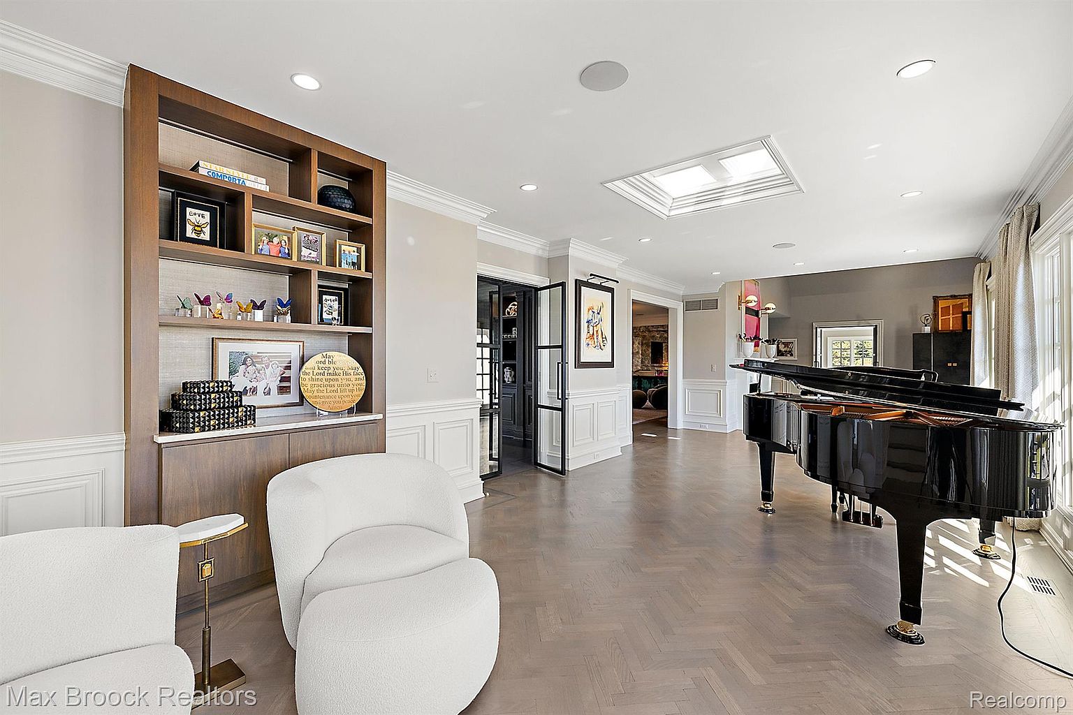 This interior shot showcases a sophisticated living room featuring a grand piano, a custom built-in bookshelf, and elegant white armchairs. The room is bathed in natural light from a skylight and large windows, highlighting the herringbone wood flooring and detailed trim work. The overall impression is one of refined luxury and comfortable elegance.