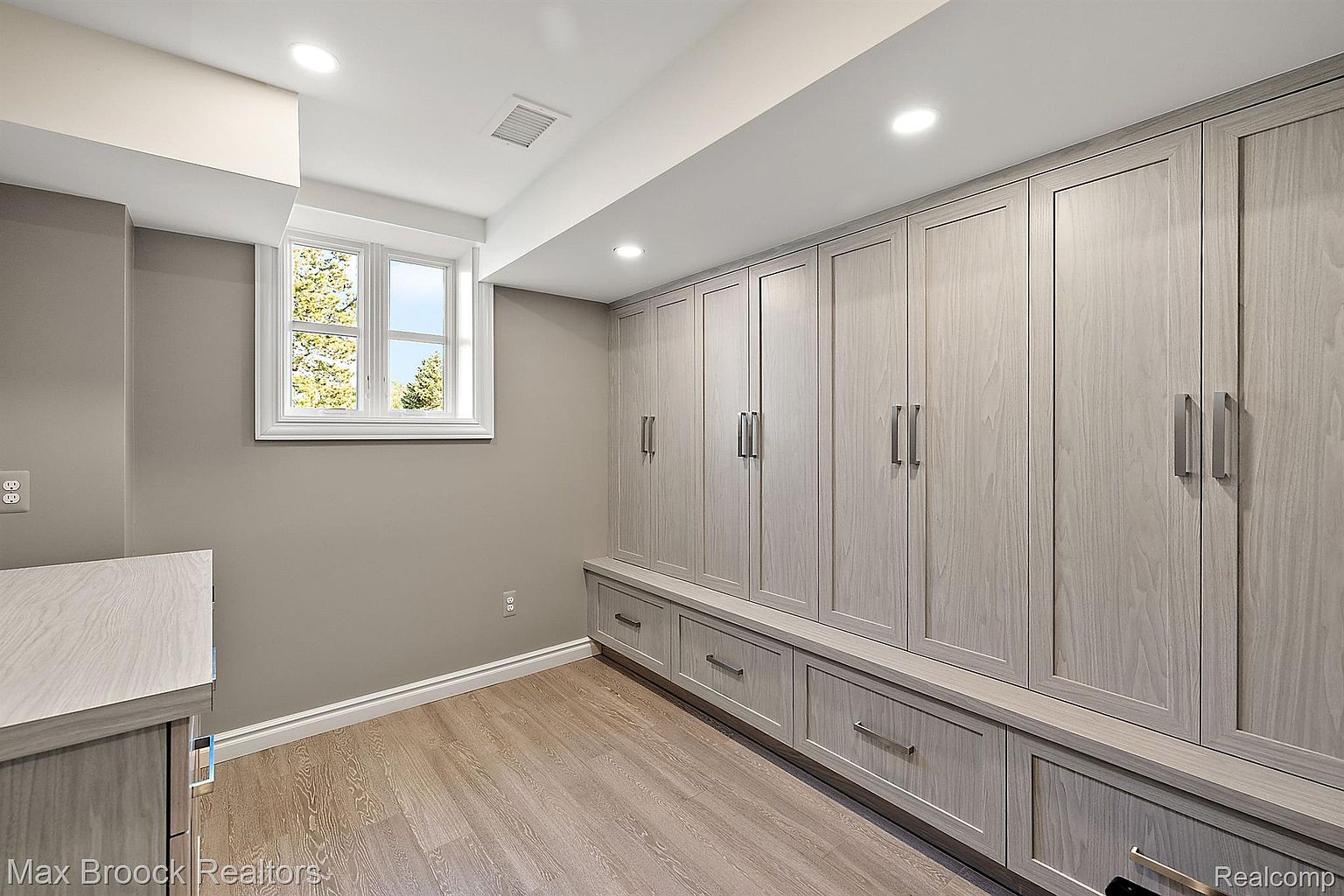 This is an interior shot of a finished basement area, featuring custom built-in cabinetry with drawers and doors in a light wood finish. A small window provides natural light, and the flooring is a light-colored wood laminate. The room has a clean and organized appearance, suggesting ample storage space.