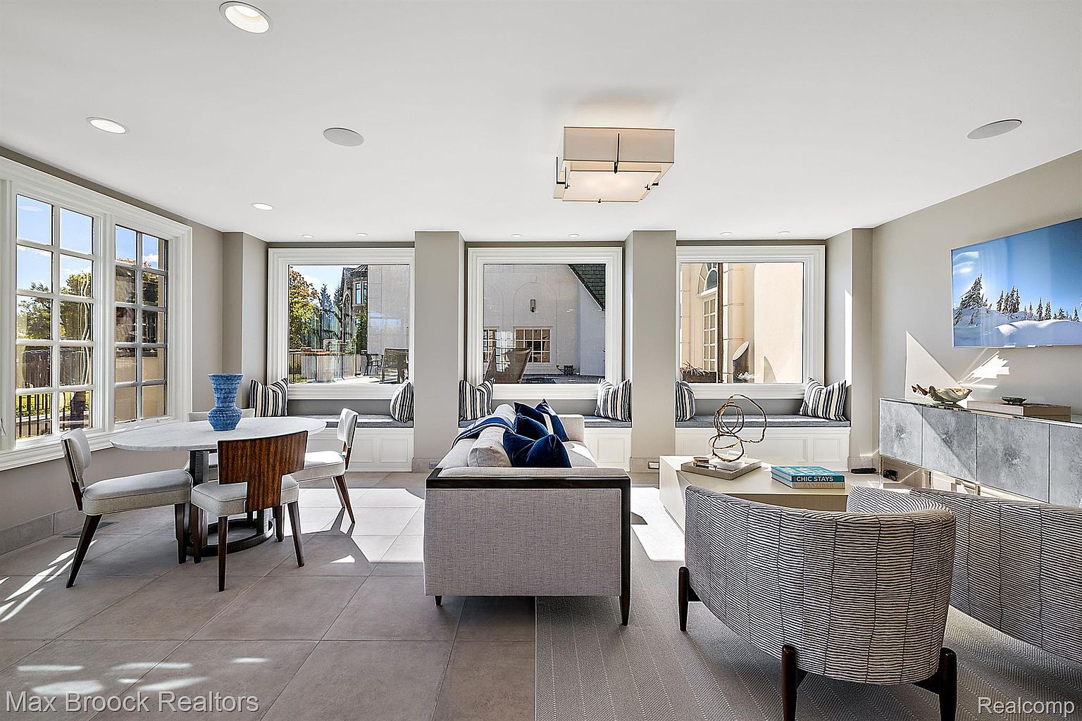 This is an interior shot of a well-lit living room, featuring a modern design with neutral tones and large windows. The room includes a dining area with a round table, a seating area with a sofa and armchairs, and built-in window seats. The overall impression is one of spaciousness and contemporary elegance.