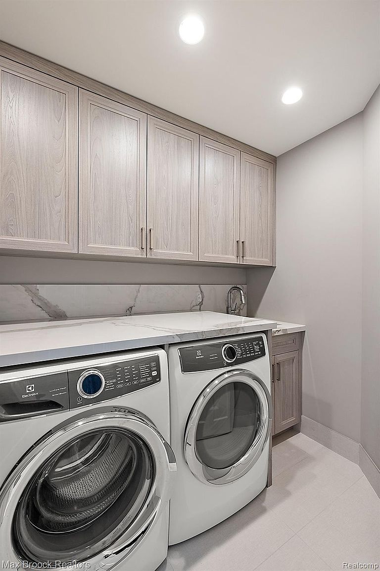 This is a well-organized laundry room featuring a front-loading washer and dryer set beneath a countertop with a marble backsplash. Above the countertop, there are light wood cabinets providing ample storage space. The room is brightly lit with recessed lighting, and the flooring appears to be light-colored tile, creating a clean and functional space.