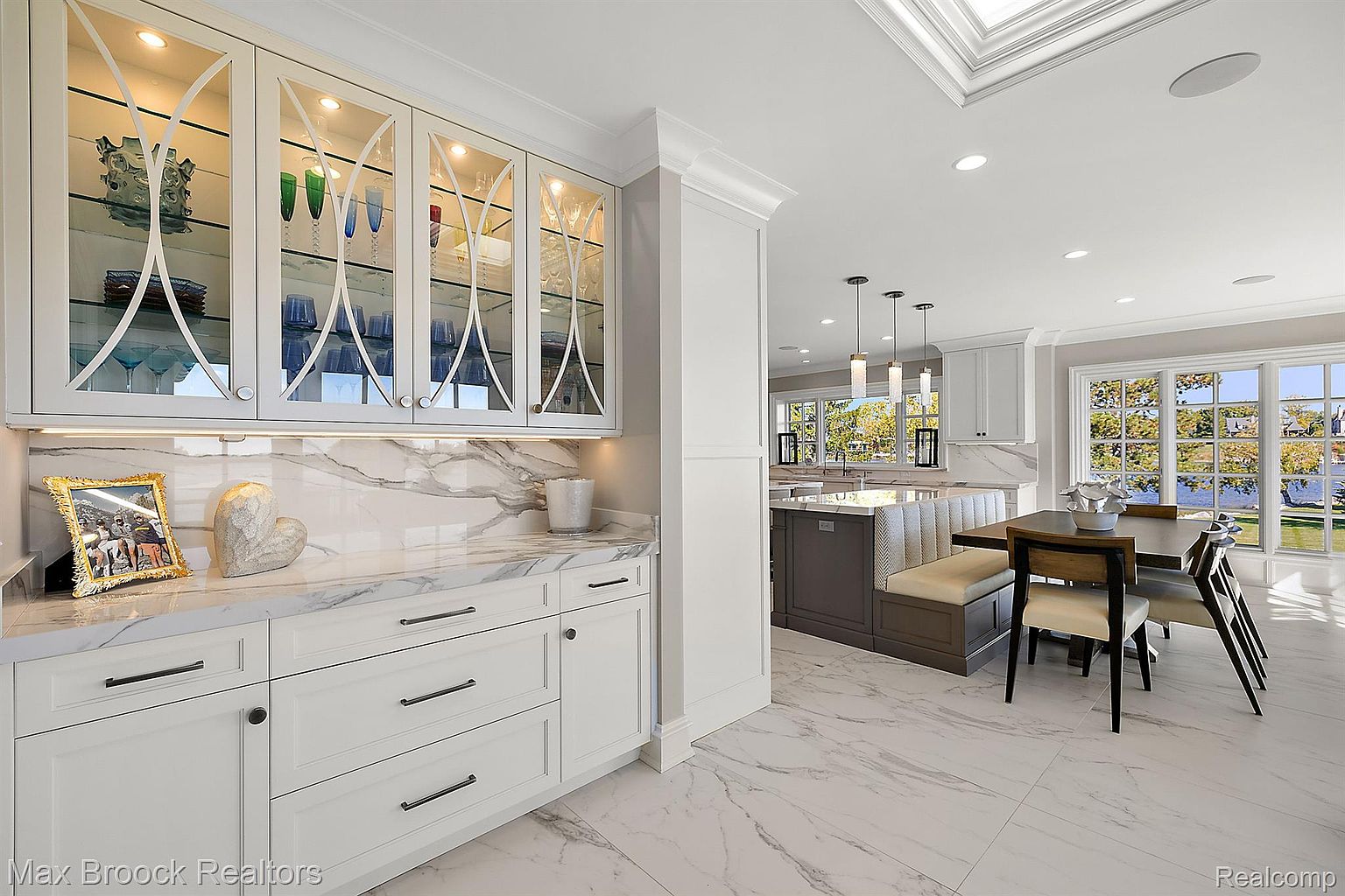 This is a bright and airy kitchen featuring white cabinetry with glass-fronted upper cabinets, marble countertops and backsplash, and marble flooring. The kitchen seamlessly transitions into a dining area with large windows offering a view of the water. The overall impression is one of luxury and sophistication.