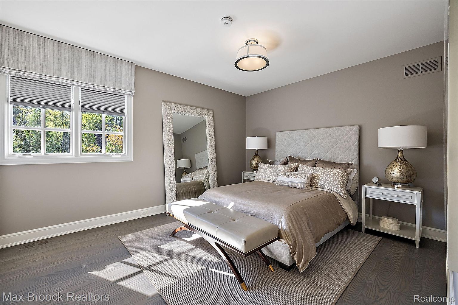 This is a well-lit primary bedroom featuring a neutral color palette with gray walls and hardwood floors. The room includes a large bed with a quilted headboard, decorative pillows, and a light-colored duvet. A full-length mirror, a white bench, and a side table with a lamp add to the room's elegance and functionality, creating a serene and inviting atmosphere.