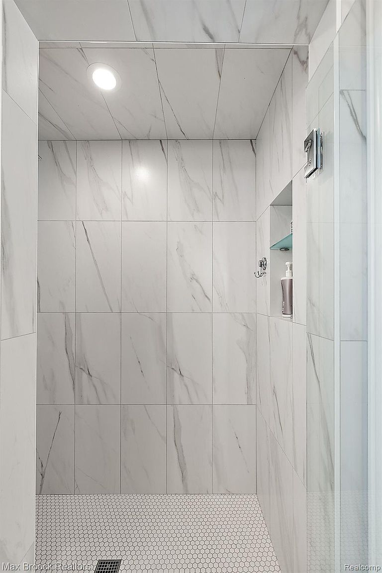 This is a well-lit bathroom featuring a shower with white marble-patterned tile walls and a white hexagonal tile floor. The shower includes a built-in niche for toiletries and a glass door. The overall impression is clean, modern, and luxurious.