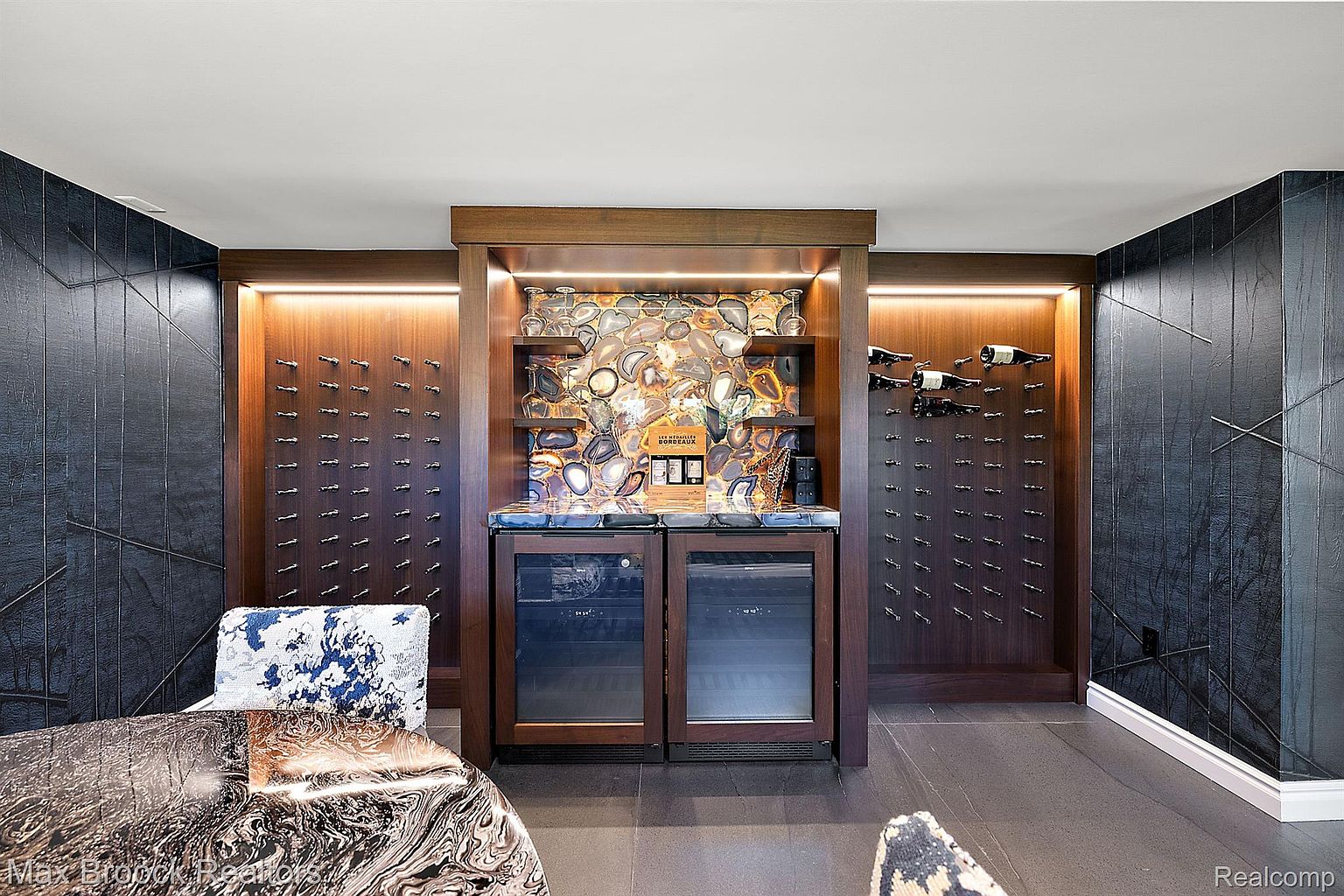 This image showcases a sophisticated wine cellar featuring custom wood cabinetry with built-in wine racks and dual wine refrigerators. A decorative stone backsplash and countertop add a touch of luxury, complemented by ambient lighting. The room exudes a modern and elegant atmosphere, perfect for wine enthusiasts.