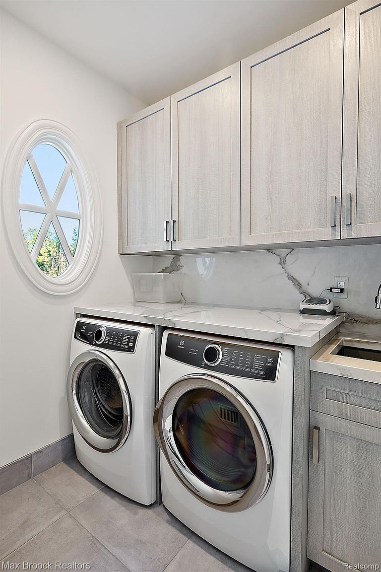 This is a well-organized laundry room featuring a front-loading washer and dryer set in white, complemented by light gray cabinetry above and to the side. A marble-patterned countertop provides ample workspace, and a round window adds natural light. The room exudes a clean and modern aesthetic, perfect for a functional yet stylish home.