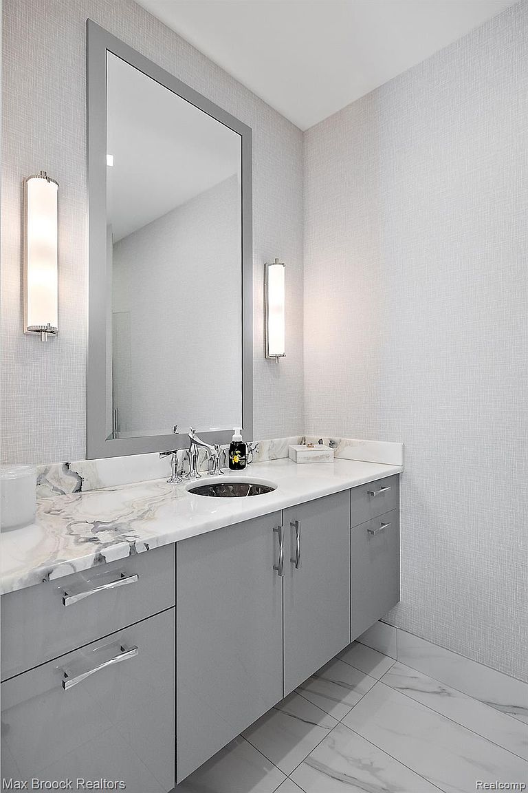 This is a well-lit guest bathroom featuring a gray vanity with a white marble countertop and a dark round sink. A large rectangular mirror is mounted above the vanity, flanked by sconce lighting. The walls are covered in a textured wallpaper, and the floor is tiled with marble.