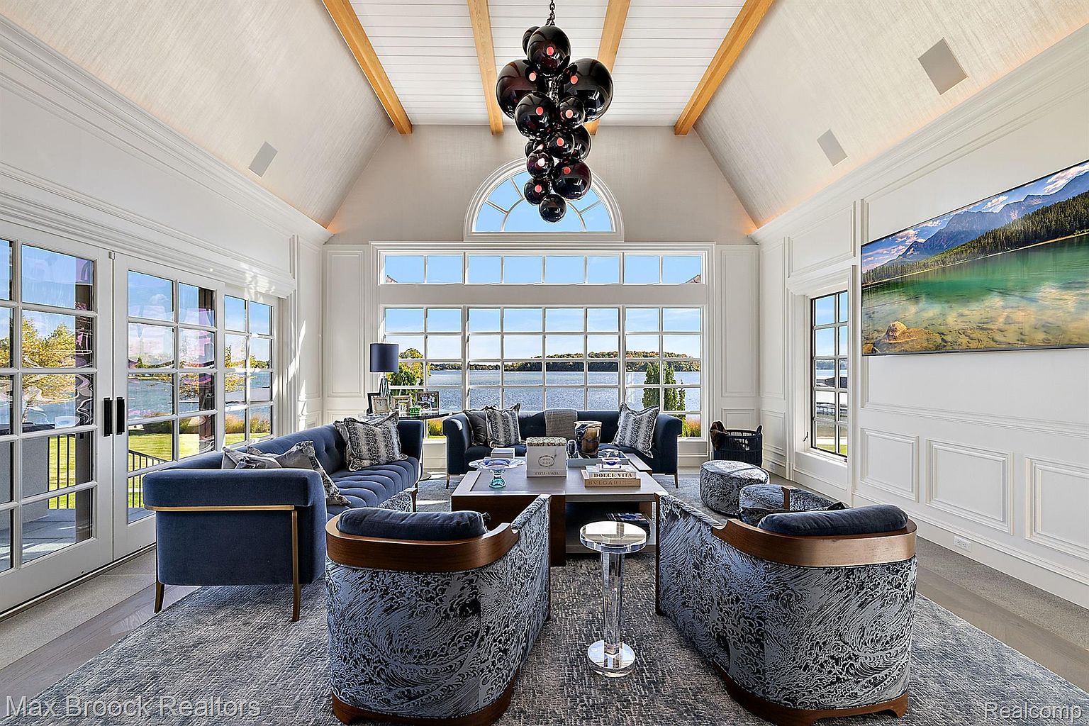 This is a luxurious living room featuring high vaulted ceilings with wooden beams and a large window offering a scenic lake view. The room is furnished with plush blue sofas, patterned armchairs, and a stylish coffee table, all set on a patterned rug. A modern chandelier hangs from the ceiling, adding to the room's elegant ambiance.