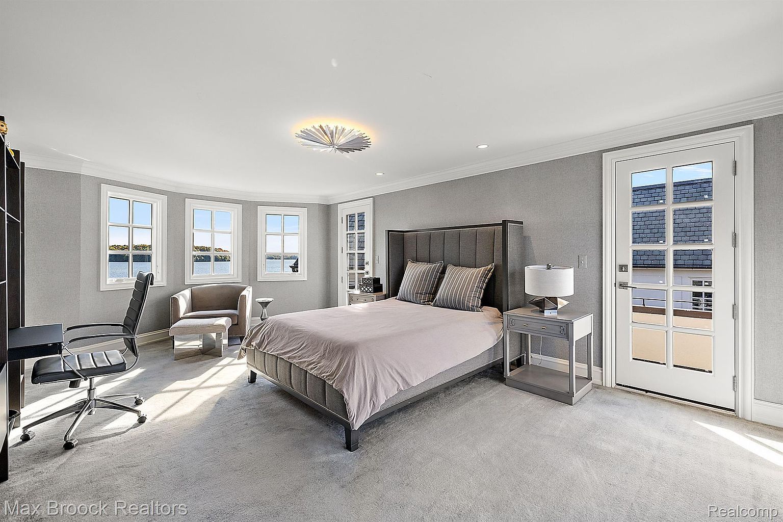 This is a primary bedroom featuring a gray color scheme with a large bed, a nightstand with a lamp, and a sitting area near the windows. The room has a modern and elegant feel, with natural light streaming in from the windows and a door leading to a balcony. The overall impression is one of comfort and sophistication.