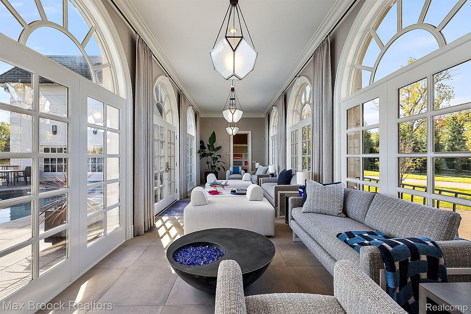 This sunroom features large arched windows and doors that flood the space with natural light, offering views of the exterior. The interior is furnished with modern sofas, a unique fire pit table, and stylish pendant lighting, creating a luxurious and inviting atmosphere. The neutral color palette and elegant design make it a perfect space for relaxation and entertaining.