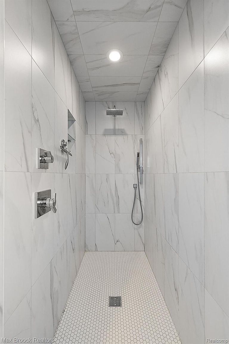 This is a modern primary bathroom featuring a walk-in shower with white marble-like tile walls and a white hexagon tile floor. The shower includes a rainfall showerhead, a handheld shower wand, and built-in shelving. Chrome fixtures and a recessed light add to the clean and luxurious feel of the space.