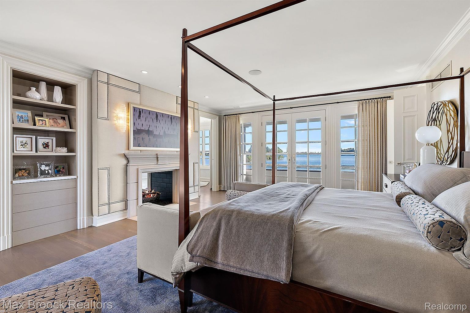 This is a luxurious primary bedroom featuring a four-poster bed with a neutral-toned duvet and decorative pillows. The room includes a fireplace with an ornate mantel, built-in shelving, and large windows offering a view of the water. The overall style is elegant and serene, creating a relaxing atmosphere.