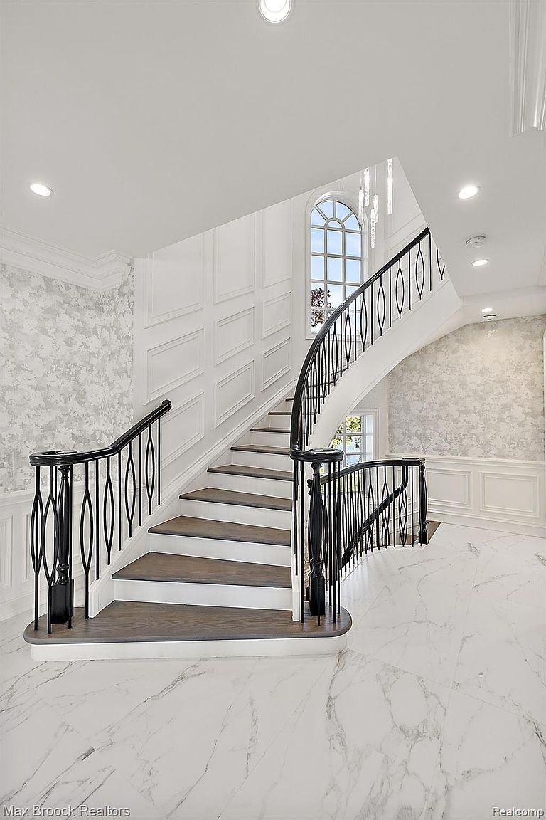 This interior shot showcases a grand staircase with dark wood treads and a black wrought iron railing. The walls are adorned with a combination of textured wallpaper and white wainscoting, complemented by a large arched window that allows natural light to flood the space. The flooring is a polished white marble, contributing to the luxurious and elegant ambiance of the home.