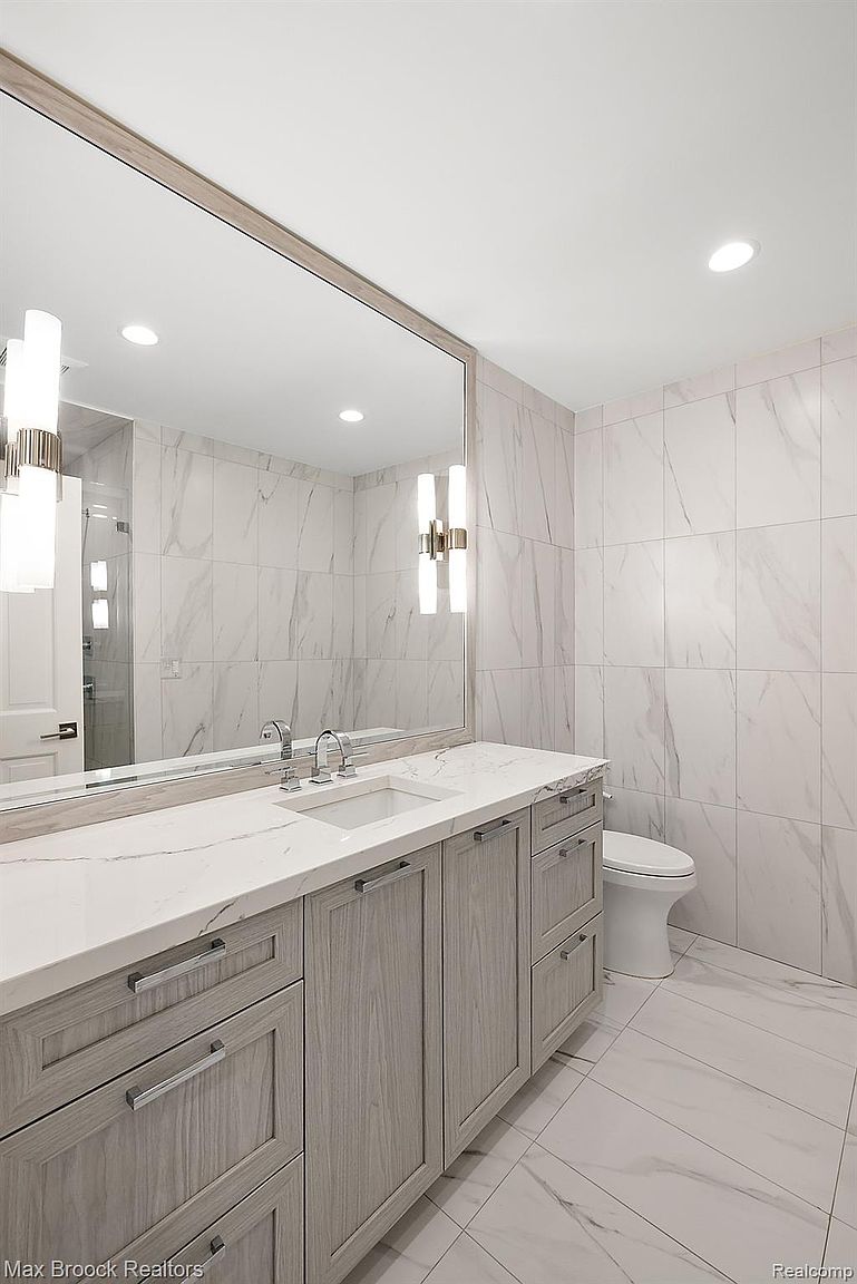 This is a well-lit primary bathroom featuring a large mirror above a vanity with gray wood-grain cabinets and a white countertop. The walls and floor are covered in white marble-patterned tiles, creating a clean and luxurious aesthetic. A toilet is visible to the right, and modern light fixtures flank the mirror.