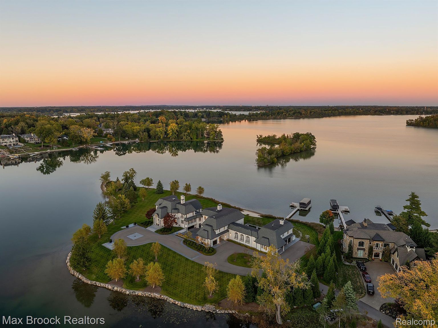 This aerial view showcases a luxurious waterfront estate at sunset. The property features a sprawling mansion with manicured lawns, mature trees, and a private dock. The surrounding lake and islands add to the property's exclusivity and scenic appeal, making it a highly desirable residence.