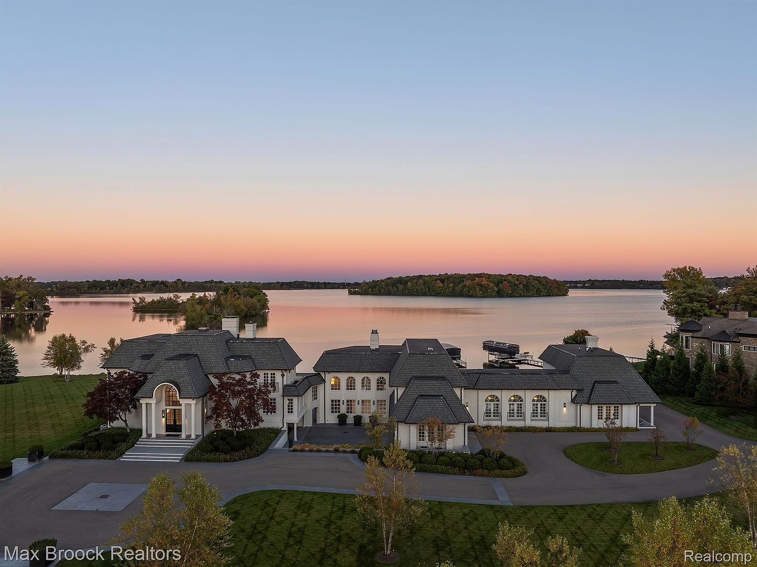 This aerial view showcases a grand waterfront estate at dusk, featuring a sprawling lawn, circular driveway, and meticulously landscaped gardens. The architecture is a blend of traditional and modern elements, with multiple wings, a dark roof, and light-colored facades. The property overlooks a serene lake with islands, creating a picturesque and luxurious setting.