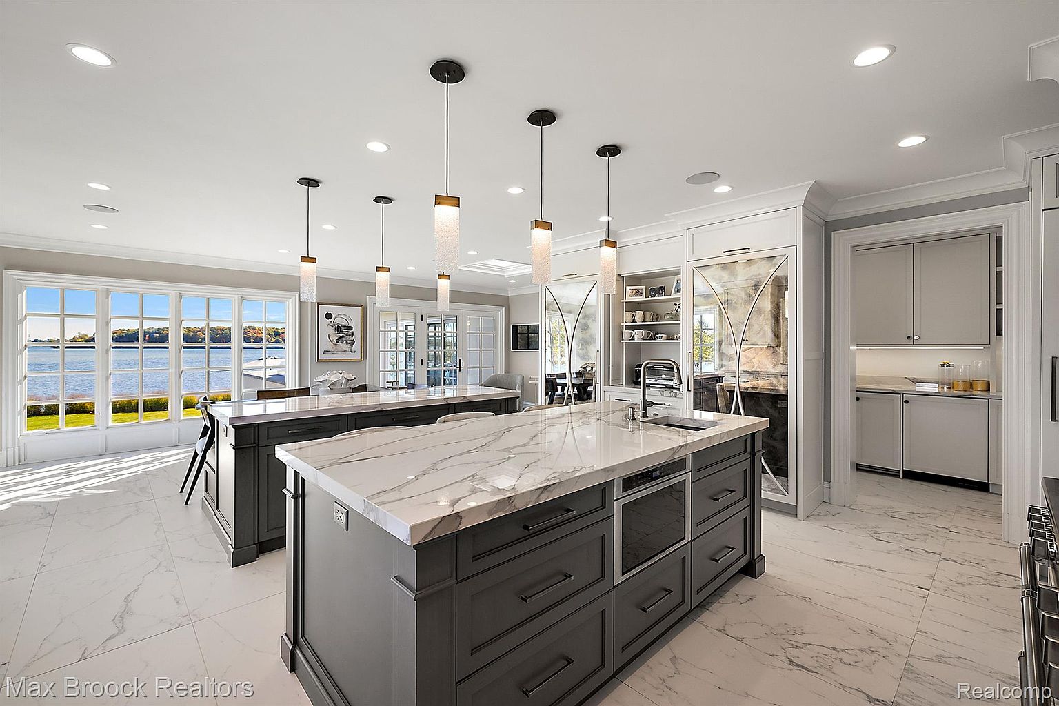 This is a bright and spacious kitchen featuring two large islands with marble countertops and dark gray cabinetry. Pendant lights hang above the islands, and large windows offer a view of the water. The kitchen also includes built-in appliances and ample storage, creating a luxurious and functional space.