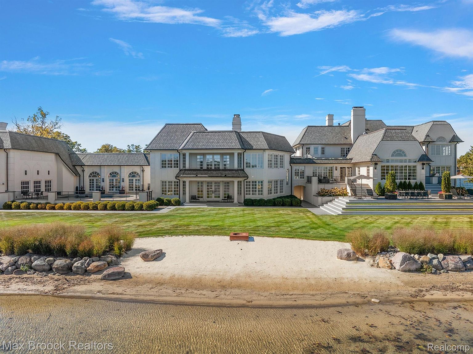 This is a rear view of a large, luxurious home with a sprawling lawn that extends to a sandy beach and waterfront. The house features multiple wings, a gray roof, and numerous windows and doors leading to outdoor spaces. The manicured lawn, beach, and water create a serene and upscale atmosphere.