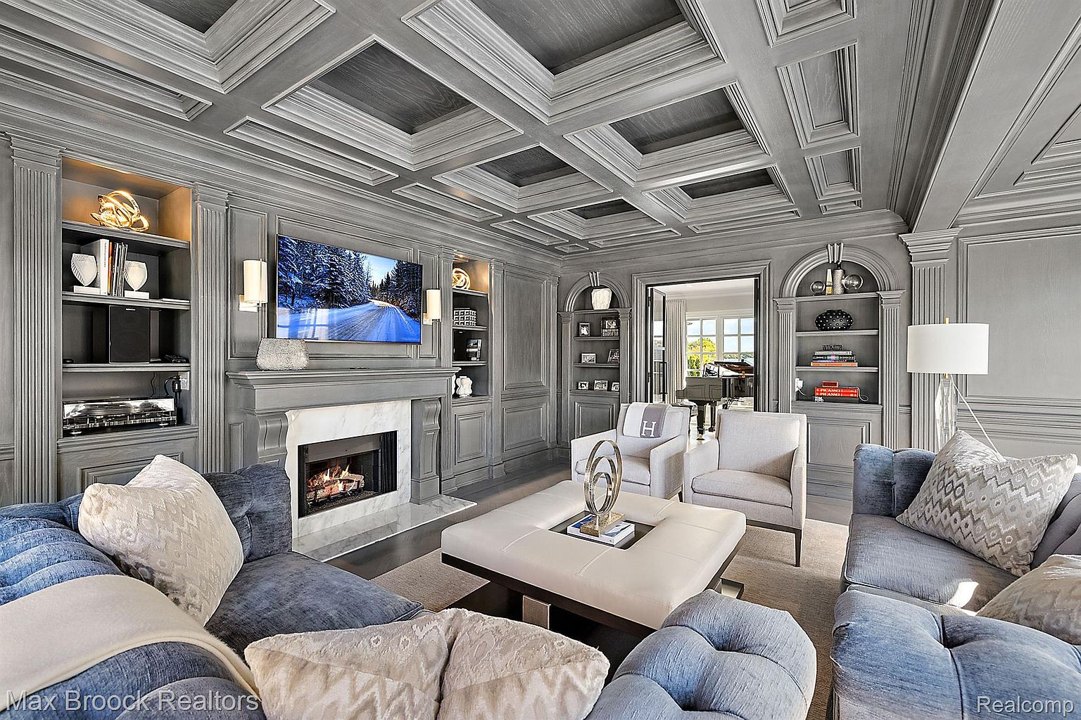This is an elegant living room featuring a coffered ceiling, custom built-in shelving, and a fireplace with a marble surround. The room is furnished with plush blue sofas, neutral armchairs, and a large ottoman, creating a comfortable and sophisticated atmosphere. A television is mounted above the fireplace, and the room is bathed in soft, natural light.