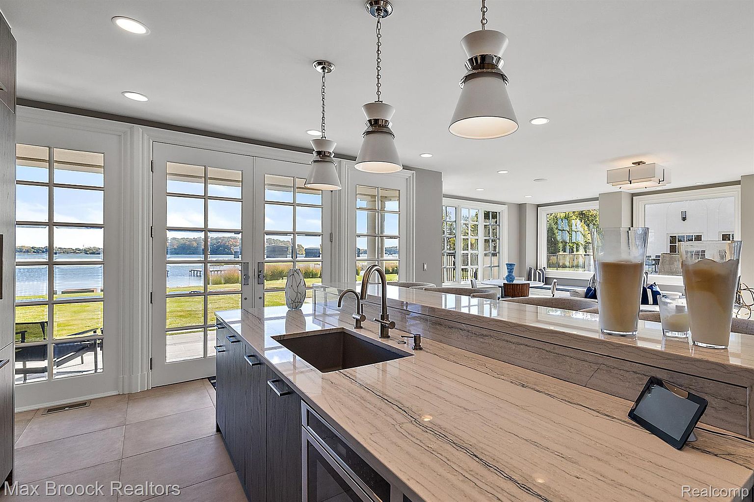 This is a bright and modern kitchen featuring a large island with a unique marble countertop and a stainless steel sink. The kitchen has dark wood cabinetry and is illuminated by pendant lighting. Large windows and glass doors offer a stunning view of the waterfront, creating a seamless connection between indoor and outdoor living.