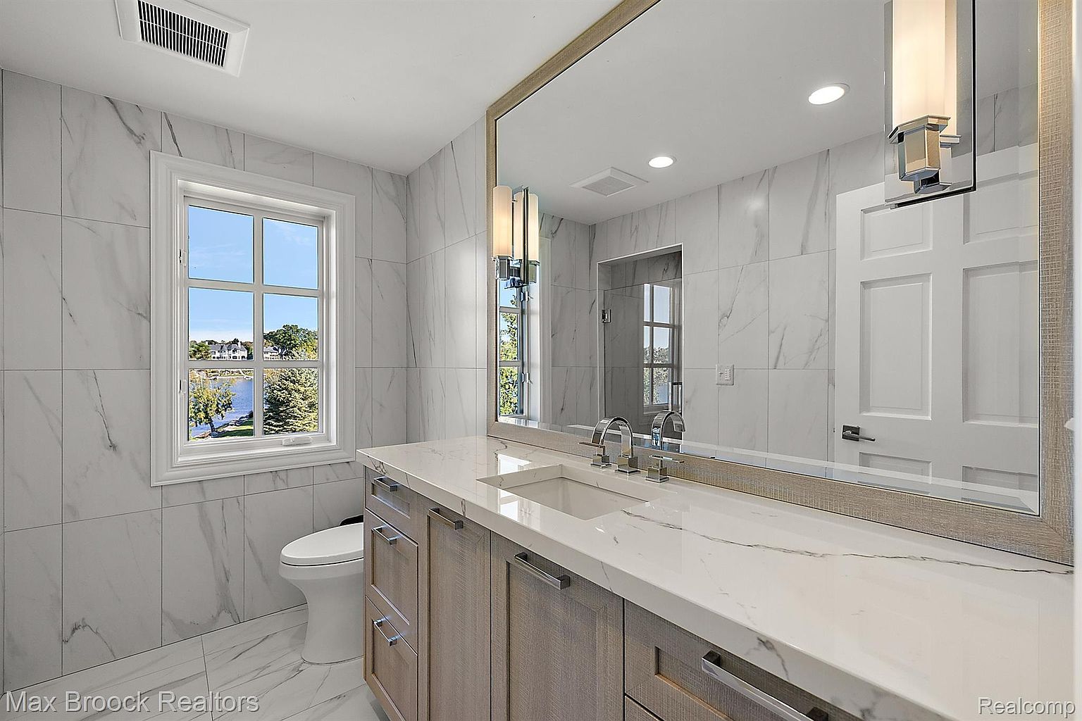 This is a primary bathroom featuring marble-like tile walls and flooring, a long vanity with light wood cabinets and a white countertop, and a large mirror. A window provides natural light and a view of the outdoors, while modern light fixtures illuminate the vanity area. The overall impression is clean, bright, and luxurious.