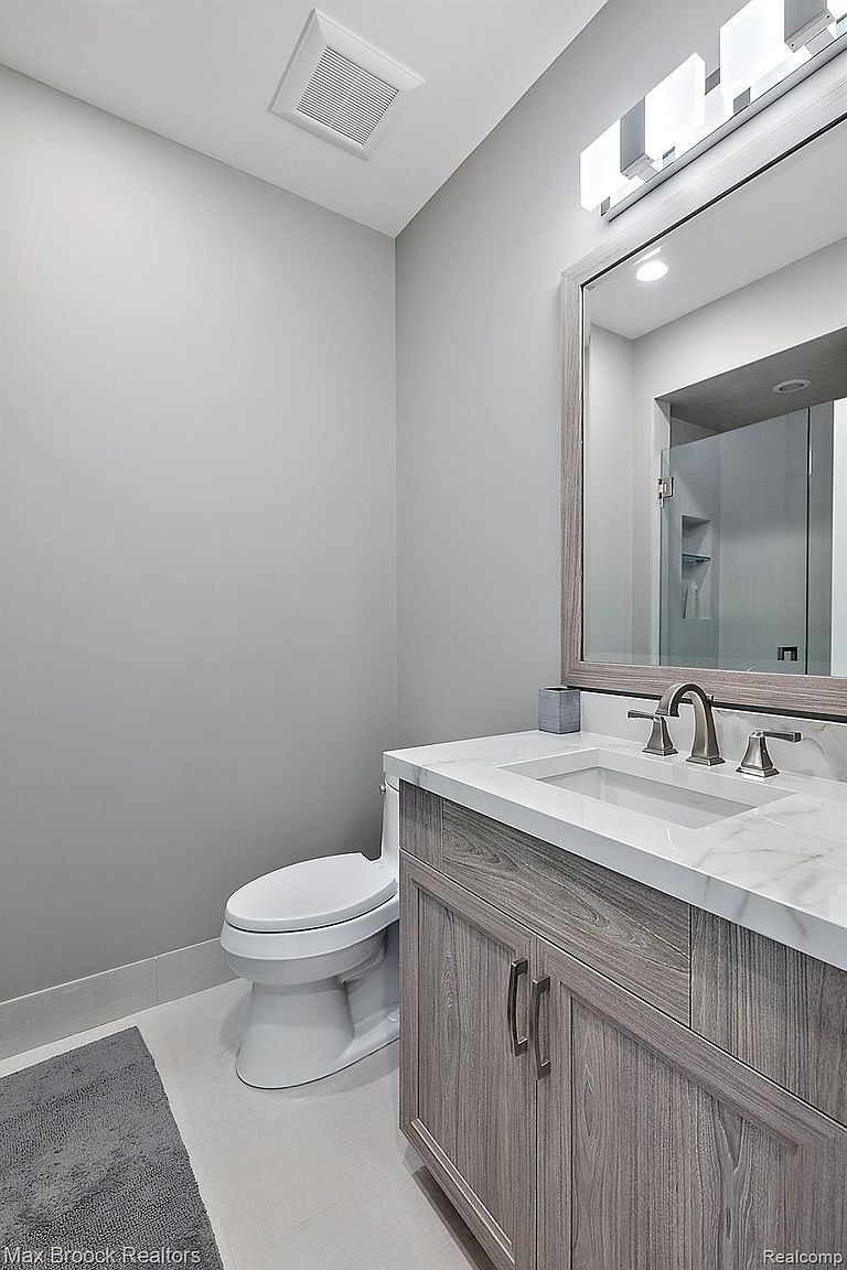 This is a well-lit guest bathroom featuring a modern vanity with a white countertop and gray wood-grain cabinets. A large mirror reflects the space, and a toilet is positioned to the left of the vanity. The walls are painted in a neutral gray tone, creating a clean and contemporary aesthetic.