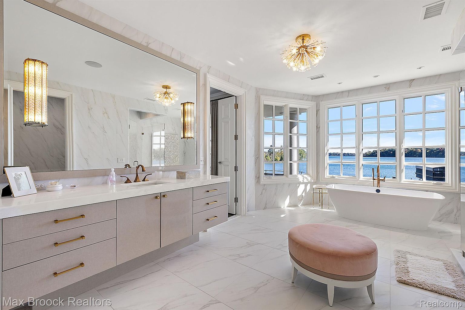 This is a luxurious primary bathroom featuring a large vanity with a marble countertop, gold hardware, and a large mirror. A freestanding bathtub sits beneath a bank of windows offering a scenic view of the water. The room is decorated in a neutral color palette with marble flooring and elegant lighting fixtures, creating a spa-like atmosphere.