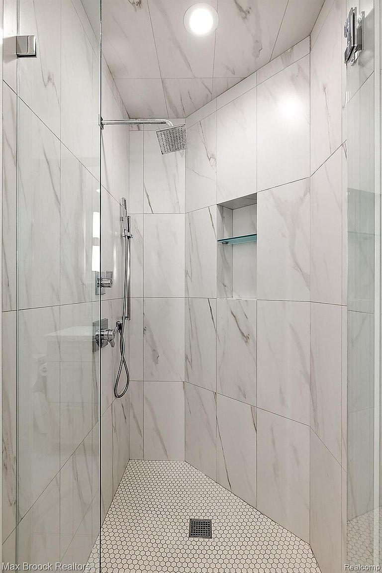 This is a well-lit primary bathroom shower featuring white marble-style tile walls and a hexagonal tile floor. The shower includes a rainfall showerhead, a handheld shower wand, and a built-in niche with a glass shelf. The overall impression is clean, modern, and luxurious.
