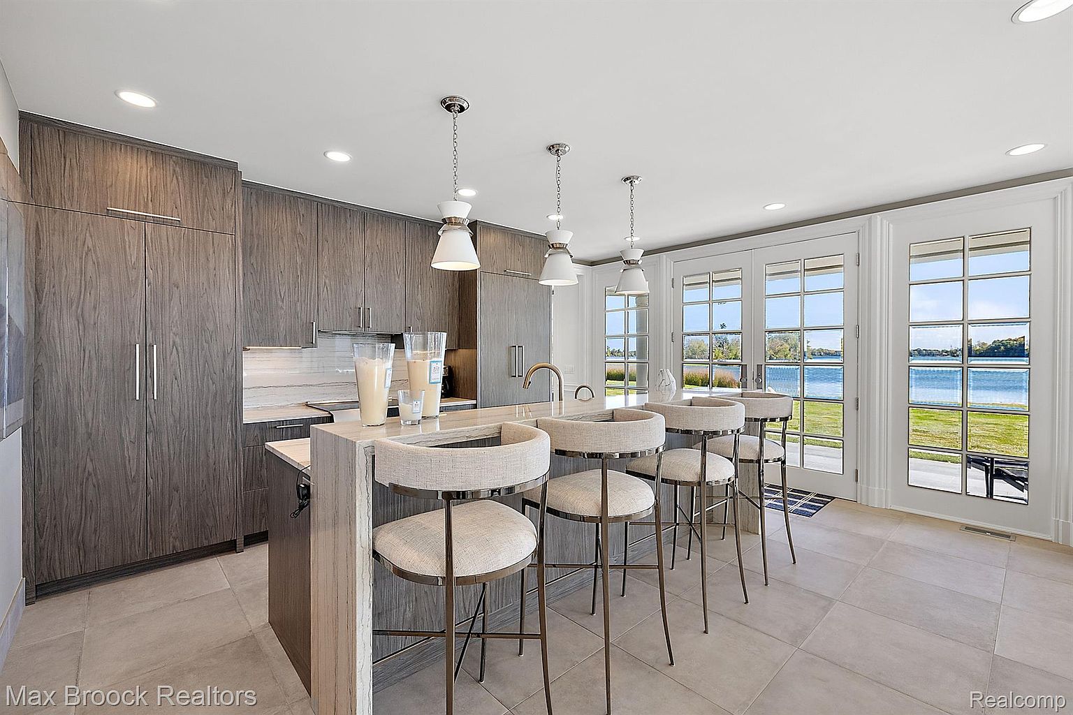 This is a bright and modern kitchen featuring sleek, wood-grain cabinetry and a large island with seating. The kitchen is well-lit with pendant lighting and recessed lights, and large windows offer a beautiful view of the water. The overall impression is one of luxury and sophistication.