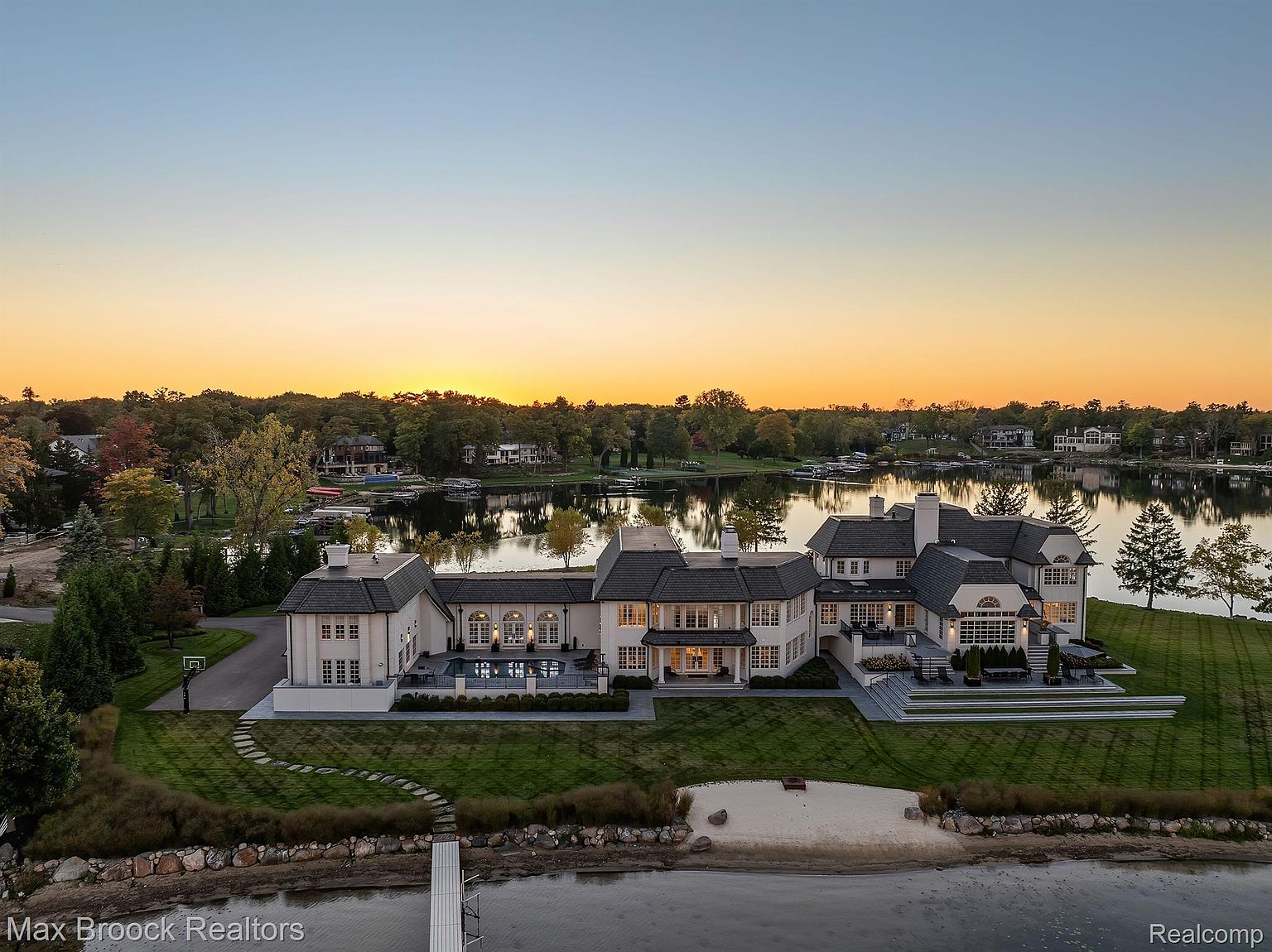 This aerial view showcases a grand estate situated on a lakefront property. The expansive home features a light-colored exterior, dark roofing, and multiple wings, complemented by a pool, patio, and private beach. Lush green lawns extend to the water's edge, creating a luxurious and serene setting.