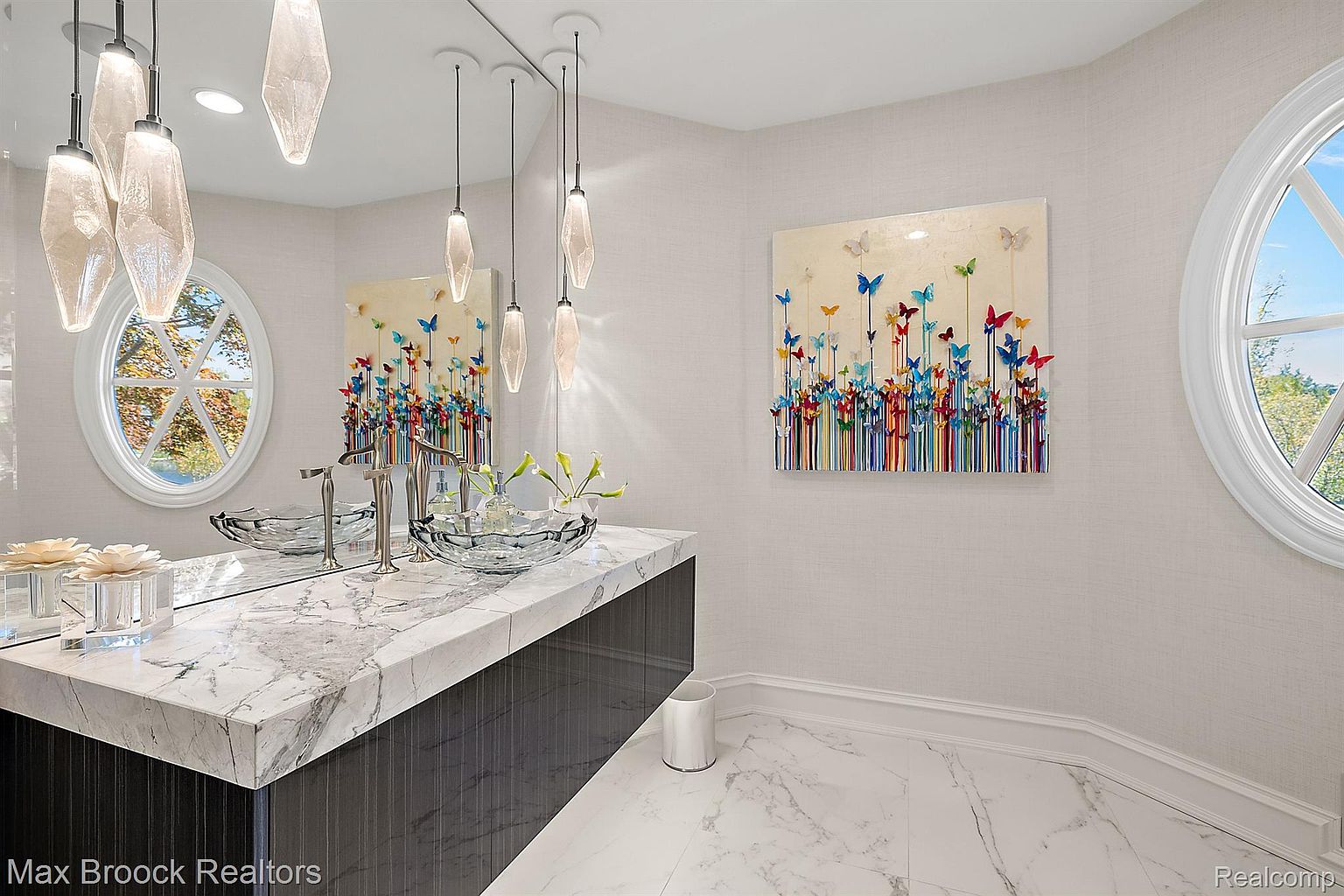 This is a stylish bathroom featuring a marble countertop vanity with a glass vessel sink and modern chrome fixtures. The walls are adorned with textured wallpaper and a colorful butterfly artwork, complemented by unique pendant lighting. A round window provides natural light, enhancing the bright and airy feel of the space, while the marble flooring adds a touch of luxury.