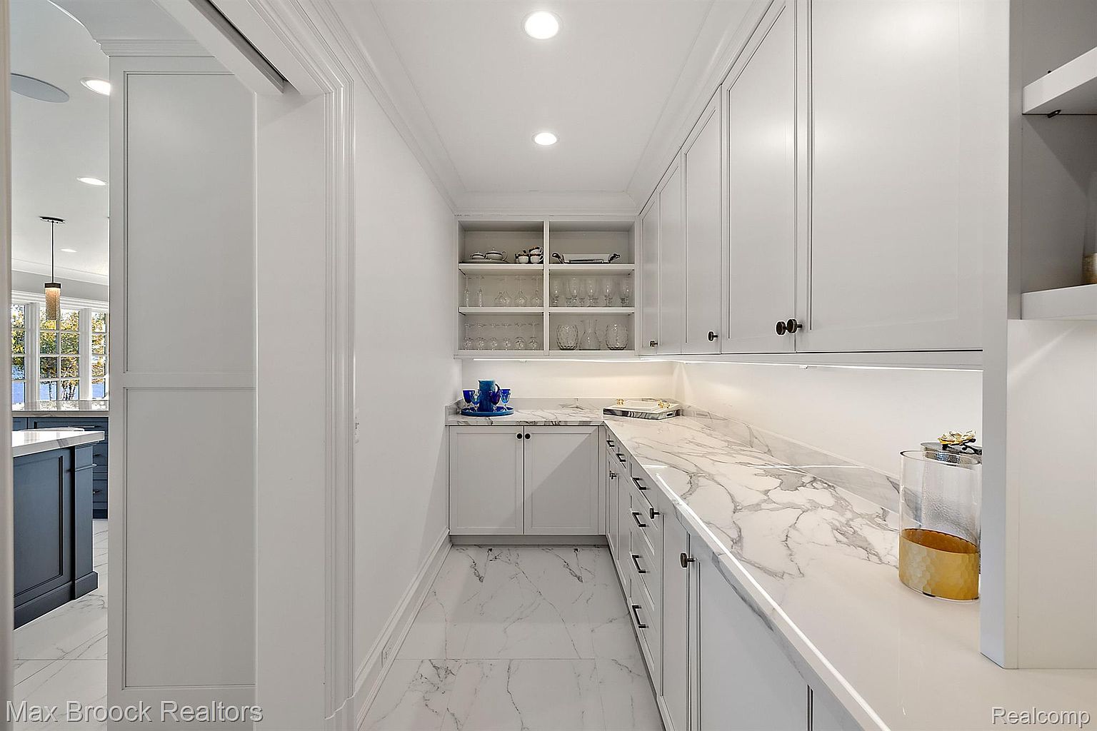 This is a well-organized pantry featuring white cabinetry, marble countertops, and open shelving. The pantry includes both closed storage below the counter and open shelving above, providing ample space for storing food and kitchenware. The marble countertops and white cabinets give the space a clean and luxurious feel.