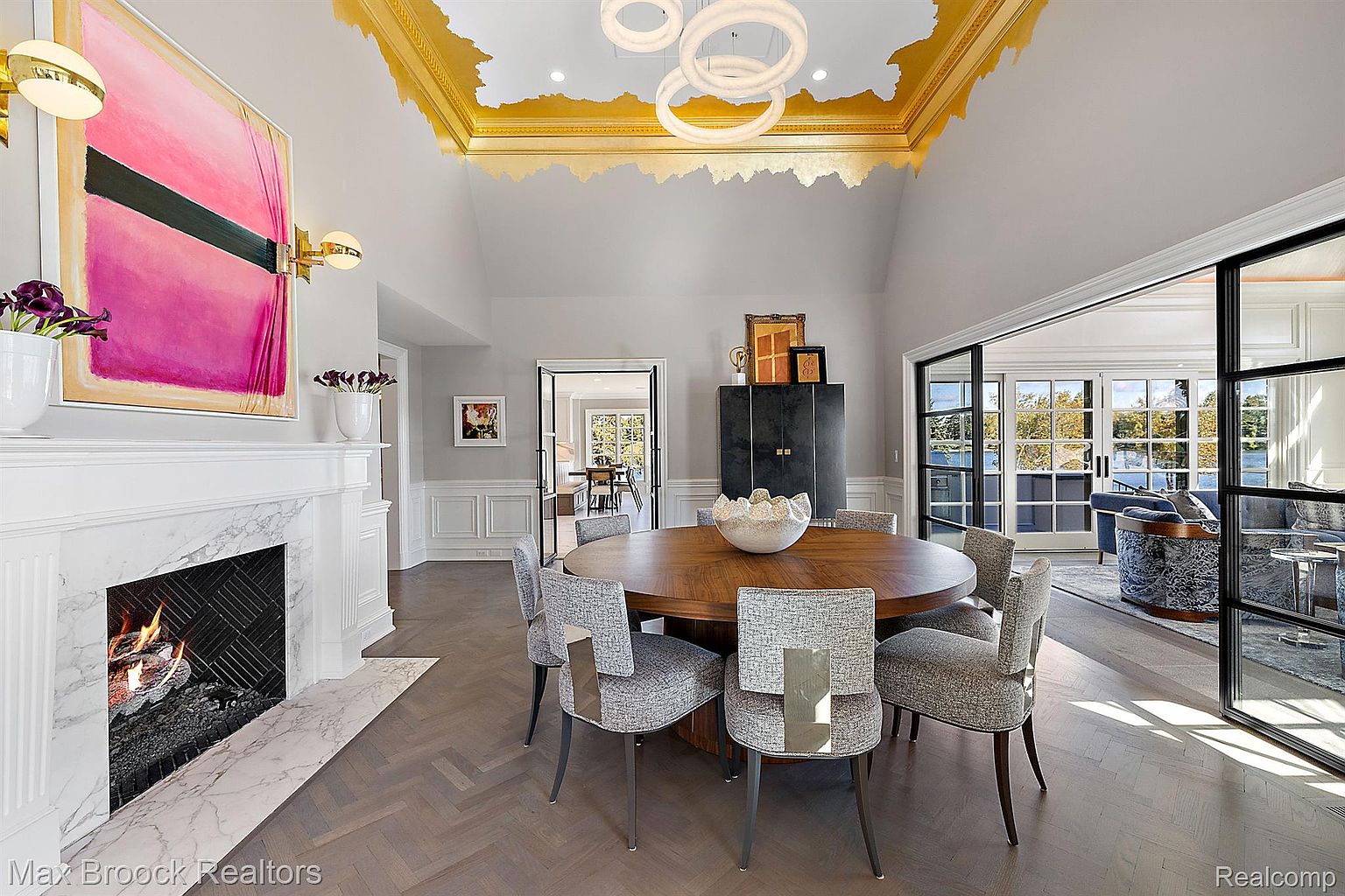 This is an interior shot of a dining room featuring a round wooden table surrounded by six upholstered chairs. A modern fireplace with a large abstract painting above it is visible on the left. The room has a high ceiling with a decorative gold leaf design and modern circular lighting fixtures. Large sliding glass doors lead to another living space, enhancing the open and airy feel of the home.