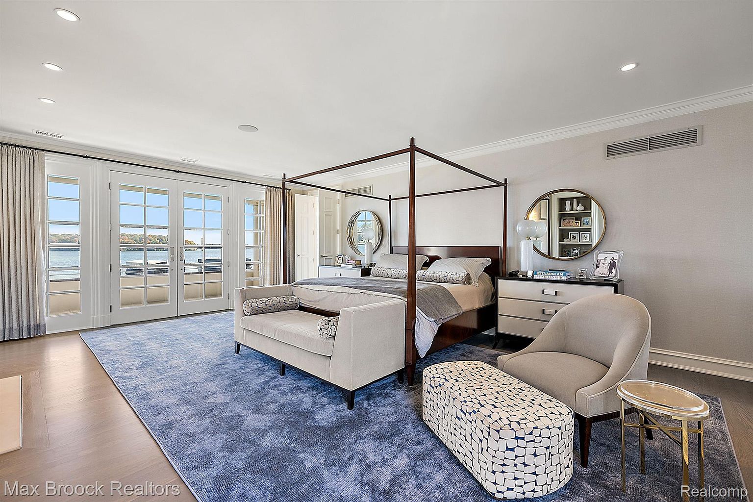 This is a spacious primary bedroom featuring a four-poster bed, a seating area with a settee and ottoman, and a view of the water through French doors. The room is decorated in neutral tones with a blue area rug, creating a serene and luxurious atmosphere. The room also features a round mirror above a dresser.